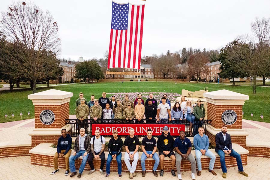 A group of Radford students and veterans sit on a sign displaying the university's name, beneath a large American flag. 