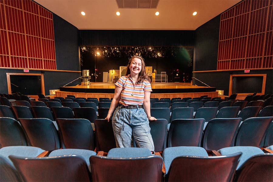 A student director sits in the seat rows of an auditorium, with her stage-setting lit behind her.