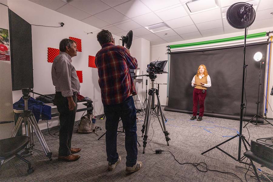 Two professors standing behind lights and a camera, recording video as a theatre student gives an advertising pitch.