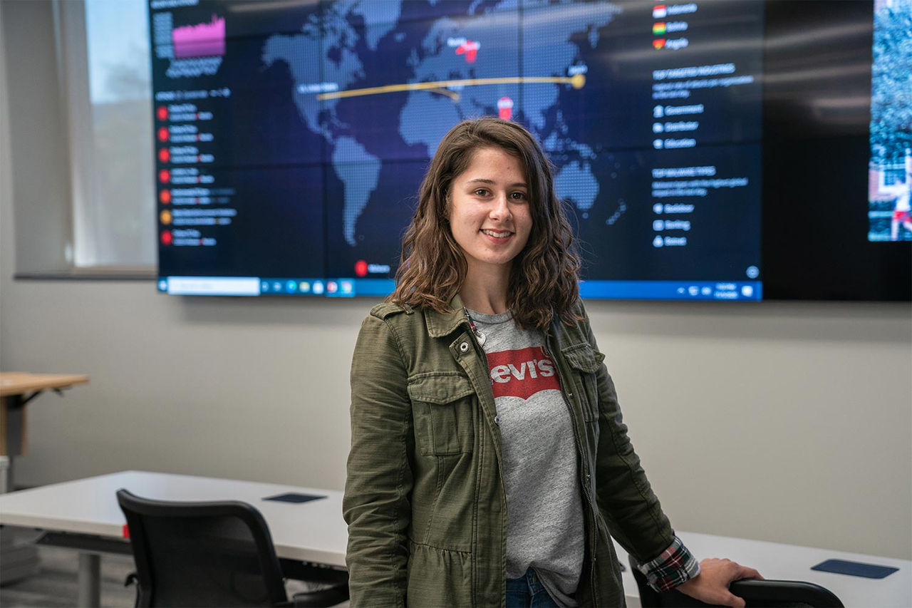 girl standing in front of a television with statistics on it smiling at the camera