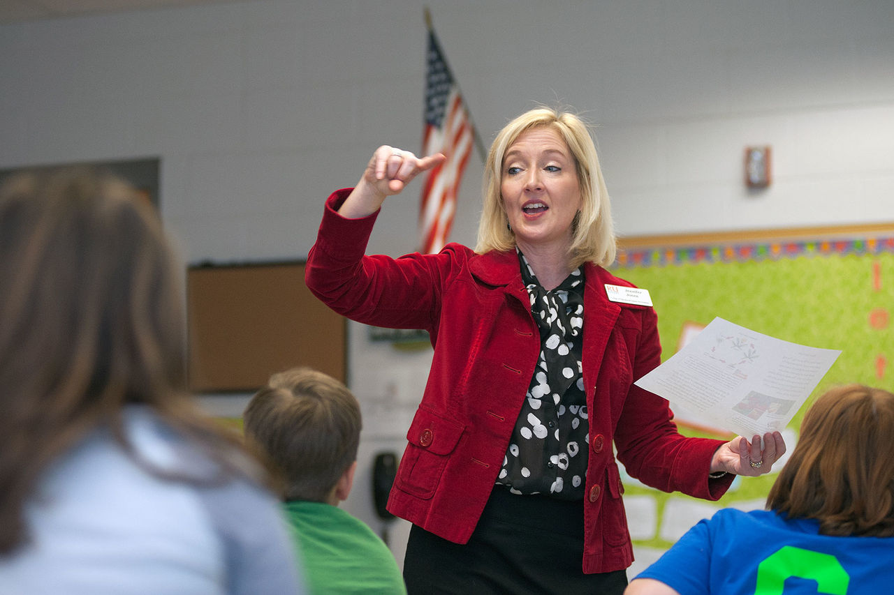 woman in a red blazer lecturing a class while holding a paper 