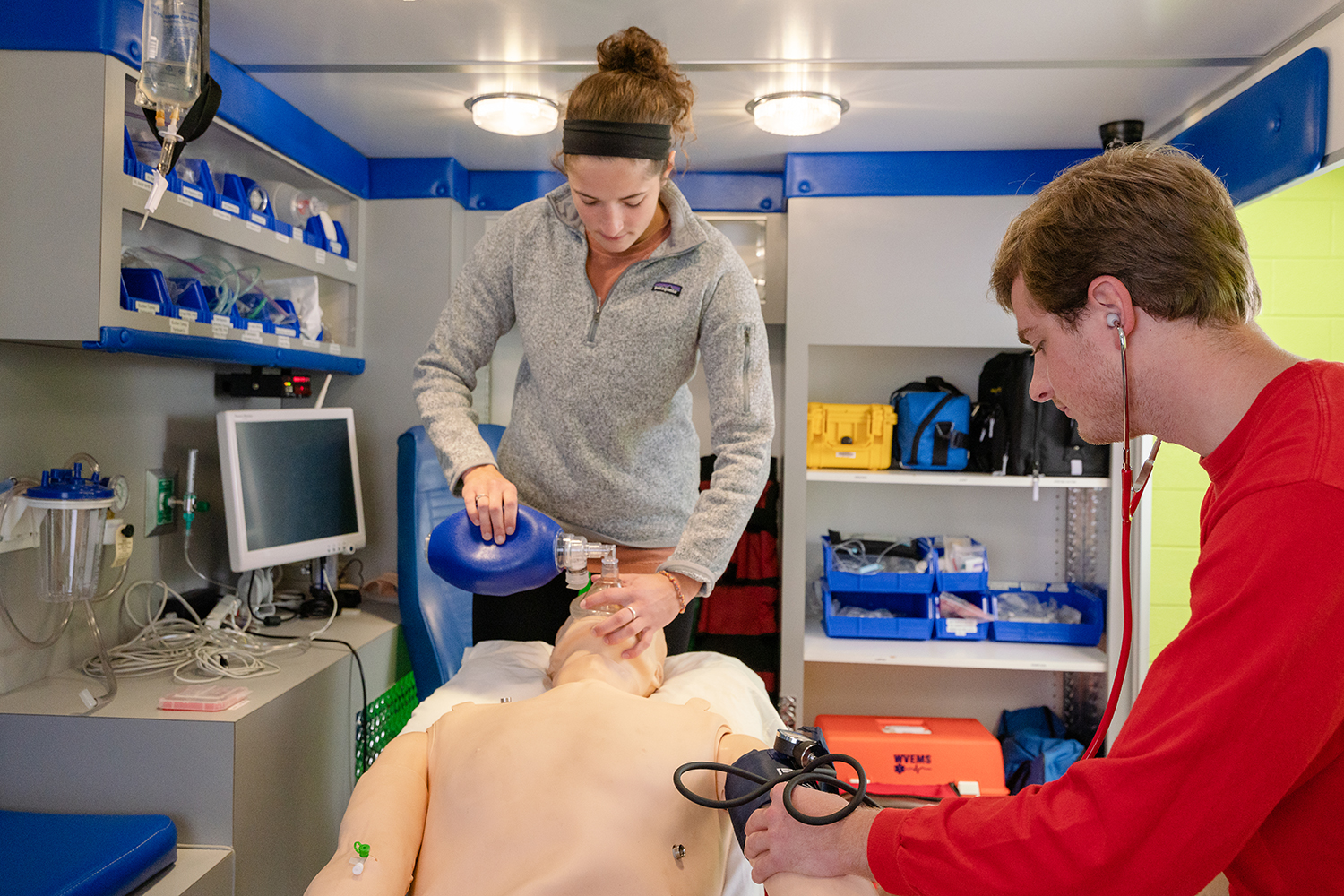 person holding a respiratory device over a mannequin while another person practices taking blood pressure