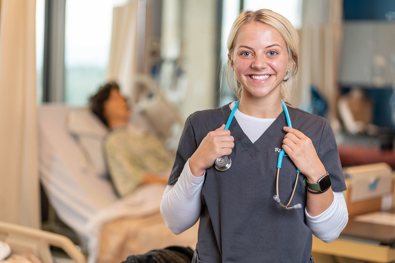 woman in scrubs and wearing a stethoscope smiling in front of a hospital bed with a mannequin patient