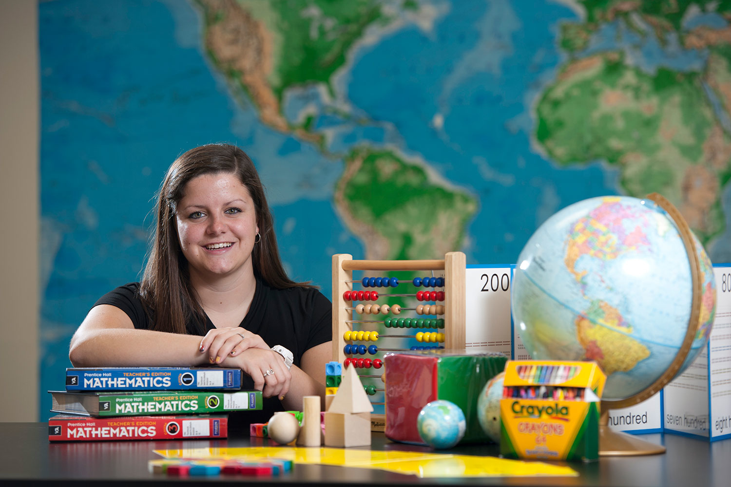 with a global map backdrop, a student sits at a desk with school supplies on it and smiles at the camera