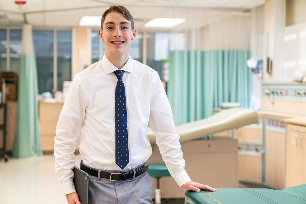 student in shirt and tie standing next to a hospital bed