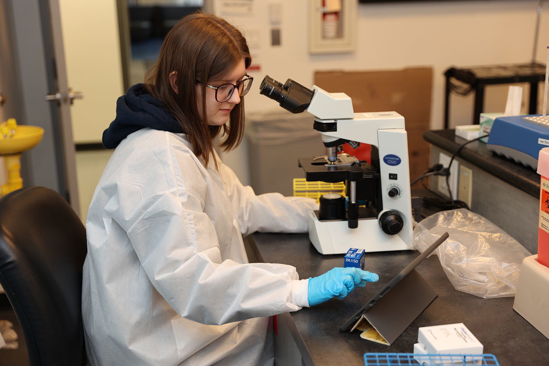 student in a lab coat looking at something on an ipad next to a microscope