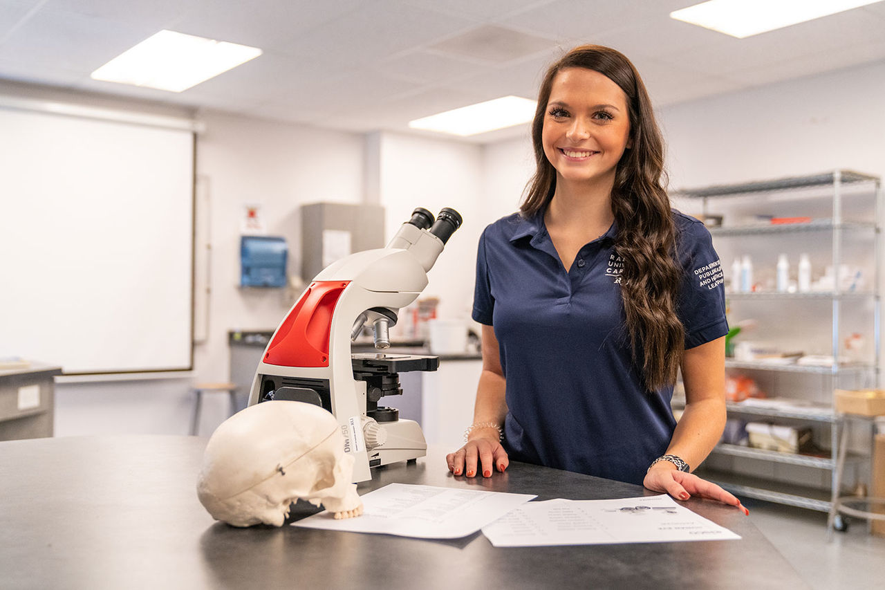 A girl in front of a desk with a microscope and a fake skill smiling at the camera