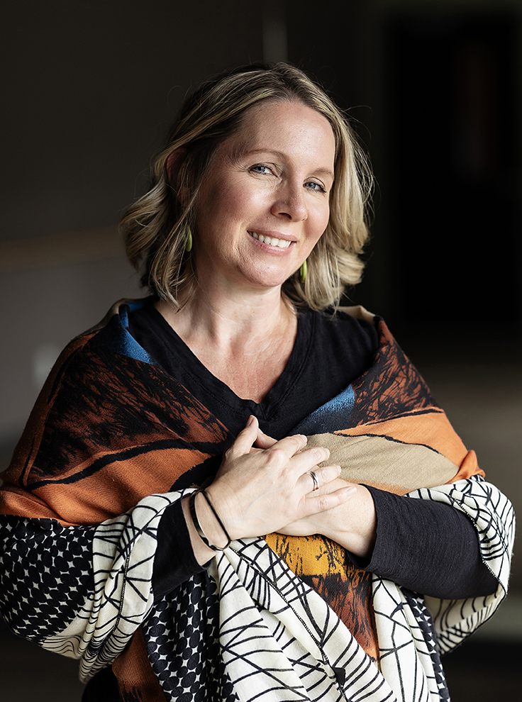 Blonde woman, Robyn Berg, in a colorful shawl against a dark background looks toward the camera.